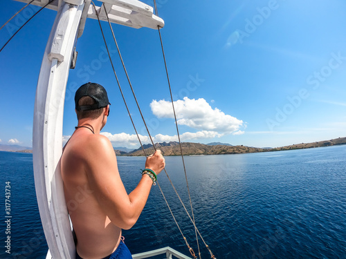 A man leaning against the mast of a sail boat enjoying beautiful island view in Komodo National Park, Flores, Indonesia. Man is enjoying the beauty of the nature. Calm sea, perfect weather for sailing