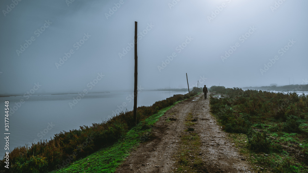 View of a dirt road with a lonely silhouette of a man walking into the distance surrounded by grassy parts and water on both sides and wooden poles on the left side, on a misty dull overcast morning