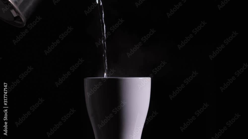Hot boiled water is poured from a metal kettle into a white ceramic cup or mug on a black background. Hot boiled water is poured from a metal kettle into a white ceramic cup or mug on a black background.