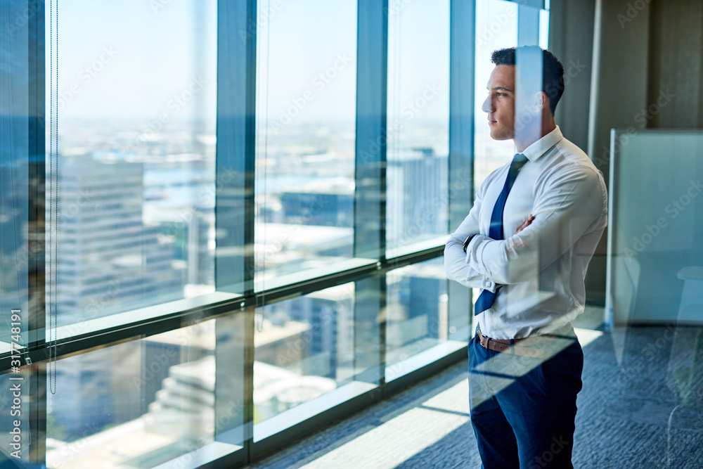 Confident young businessman deep in thought while looking through windows at the city from high up in an office building Confident young businessman deep in thought while looking through windows at the city from high up in an office building