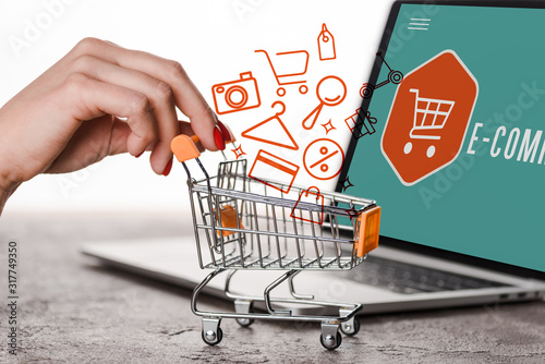 cropped view of woman holding toy shopping cart near laptop and illustration on white, e-commerce concept photo