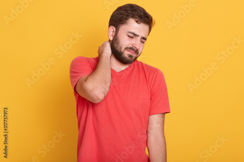 Image of attractive bearded male feeling pain in neck, frustrated young man wearing red casual t shirt, touching his neck and expressing negativity while standing against yellow studio background.