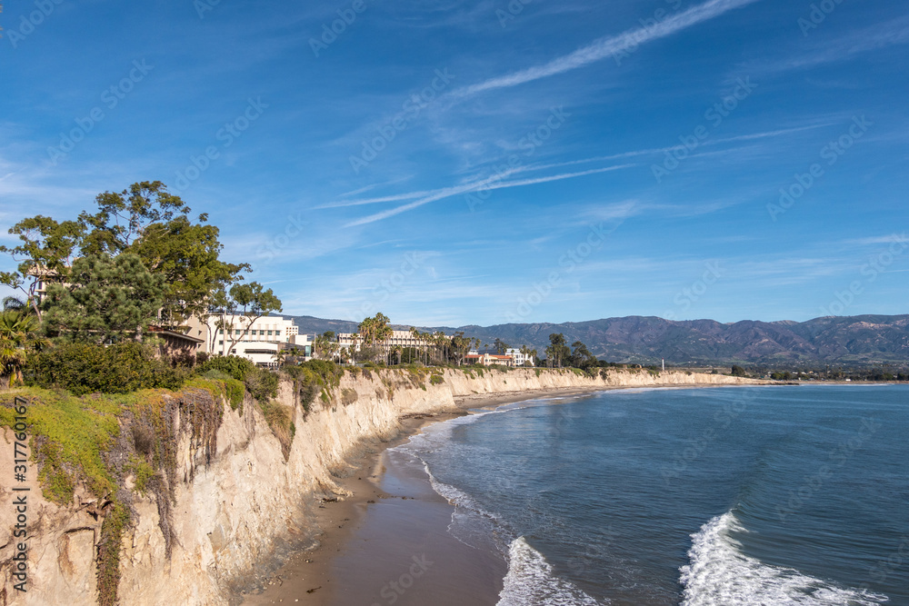 Goleta, CA, USA - January 2, 2020: UCSB, University California Santa Barbara. East side beige cliffs with green foliage bordering blue sea. Buildings in back. Hills on horizon. Blue sky.