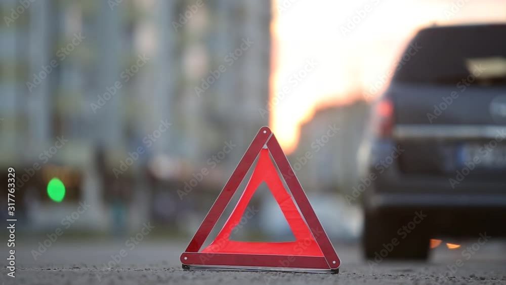 Red emergency stop sign behind broken blurred car with blinking lights on evening city street curb.