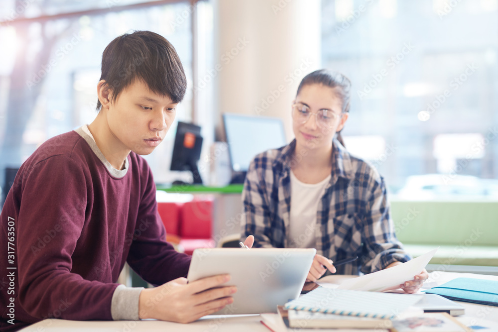 Asian young man studying and using digital tablet while sitting at the table together with young woman Asian young man studying and using digital tablet while sitting at the table together with young woman