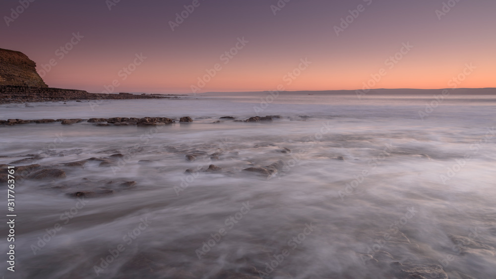 Sea water, washing over a rocky shore, while the sun sets on the horizon. A long exposure has created a milky sea
