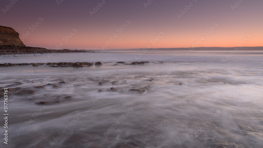 Obraz premium Sea water, washing over a rocky shore, while the sun sets on the horizon. A long exposure has created a milky sea