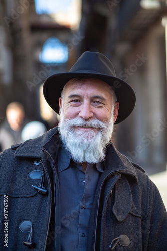 old man with white beard and black hat smiling in the middle of the street