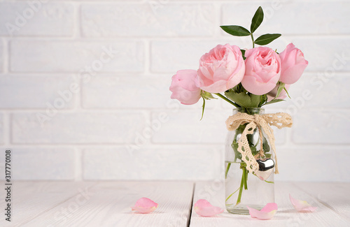 Fototapeta Naklejka Na Ścianę i Meble -  Bouquet of pink roses in a transparent glass vase with a ribbon and a metal heart on a white background with scattered petals on the table, side view, composition for holidays, weddings, postcards.