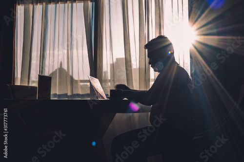 The silhouette of a man at work using a laptop in a modern room with a window background illuminated by slightly dazzling natural sun glare.
