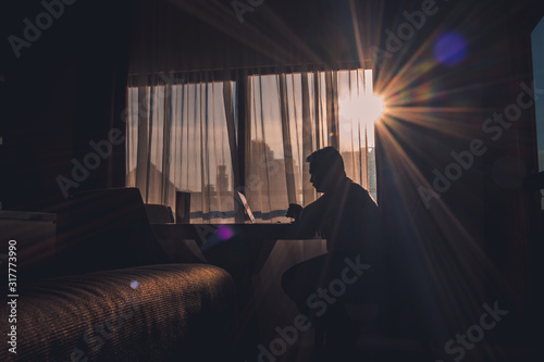 The silhouette of a man at work using a laptop in a modern room with a window background illuminated by slightly dazzling natural sun glare.