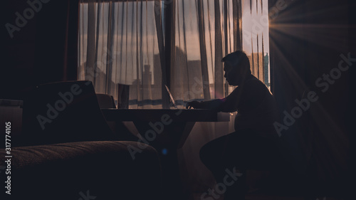 The silhouette of a man at work using a laptop in a modern room with a window background illuminated by slightly dazzling natural sun glare.