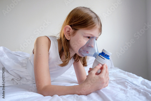 a little sick girl in white clothes sits on a white bed and breathes through an inhaler mask to treat a throat