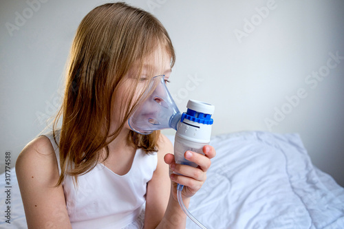 a little sick girl in white clothes sits on a white bed and breathes through an inhaler mask to treat a throat