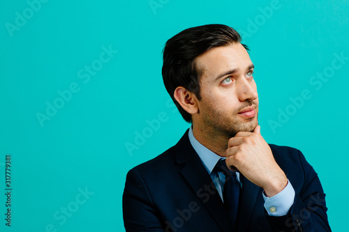 Portrait of a young entrepreneur business man thinking with hand under his chin, isolated on blue studio background