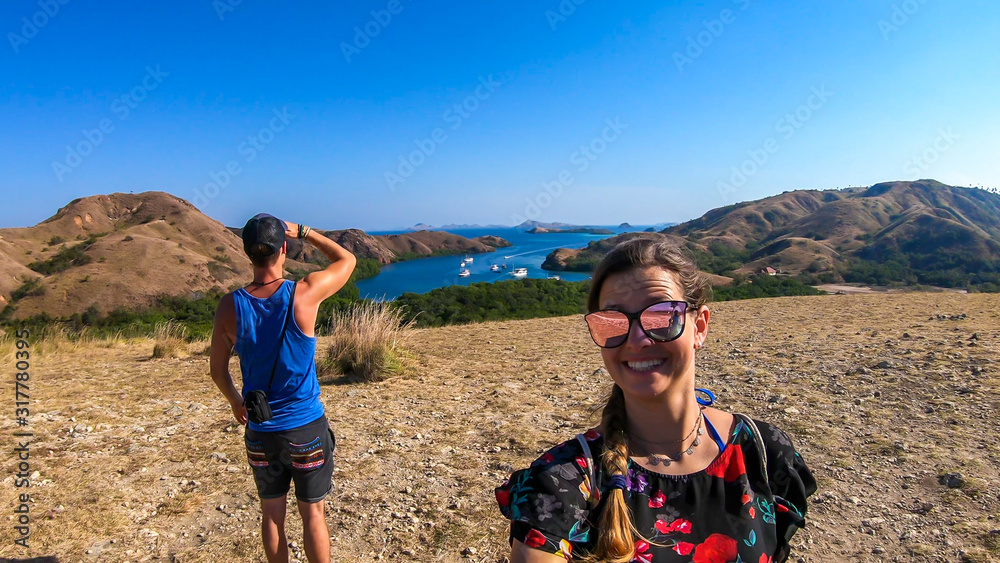 A couple standing on top of the Komodo Island in Indonesia. The man is looking at the bay, the woman smiles to the camera. Happiness and fun while travelling. Discovering new places. A couple standing on top of the Komodo Island in Indonesia. The man is looking at the bay, the woman smiles to the camera. Happiness and fun while travelling. Discovering new places.
