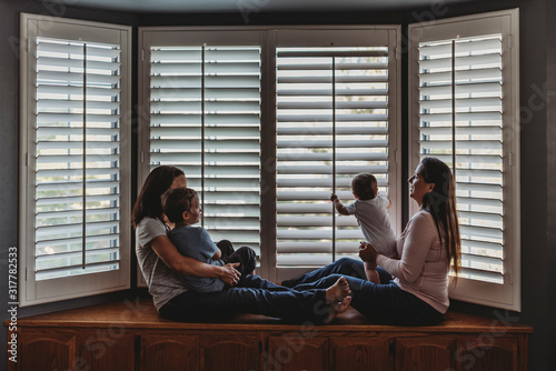 Same sex parents lounging with kids on window seat with tall shutters photo