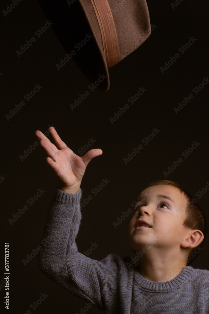 Studio portrait of boy in a gray sweater, on a black background, tossing up a brown hat with his hands Studio portrait of boy in a gray sweater, on a black background, tossing up a brown hat with his hands