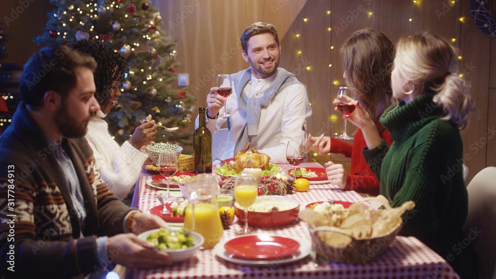 Happy multi-racial attractive friends toasting with red wine at Christmas table eating dinner celebrating holidays. Noel evening. Young people party.