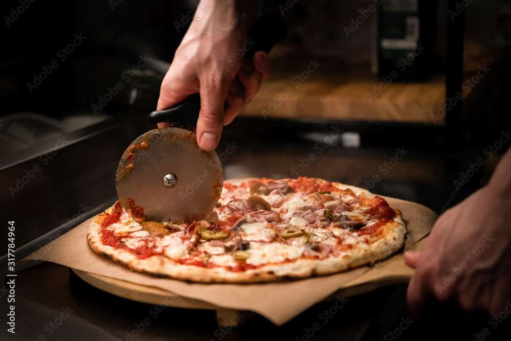 Chef cutting freshly baked delicious pizza with pizza cutter on professional kitchen. Close up of a man cutting pizza on wooden board. Traditional italian food restaurant. Low key, hand's close up