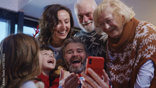 Happy extended family with grandparents making a fun self portrait together using mobile phone embracing together celebrating Christmas. Noel gathering.