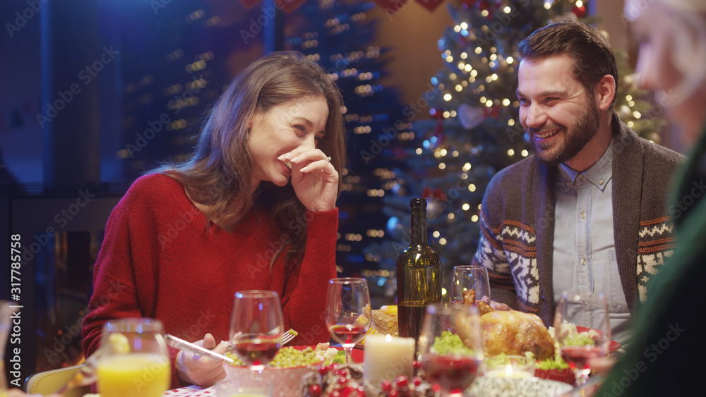 Attractive cheerful caucasian couple of man and woman sitting and laughing at dining table celebrating Christmas holiday dinner with friends.