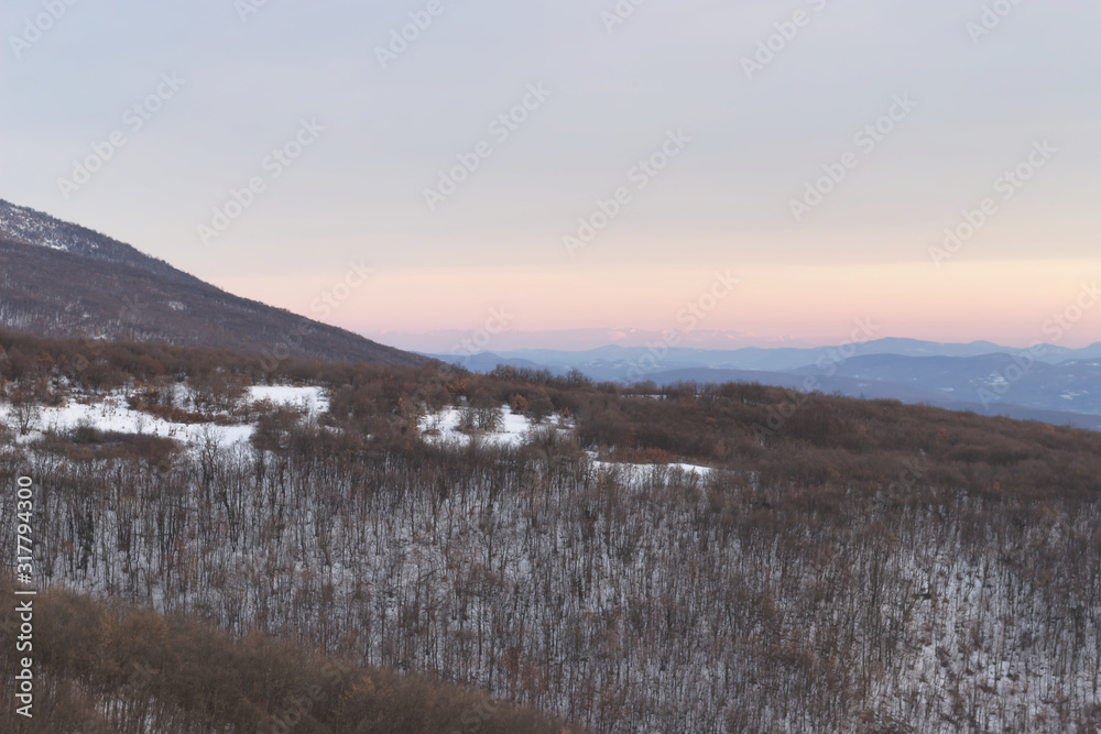 Panoramic view of a forest on a snow covered mountain with distant mountain layers and soft colors of a sunset sky Panoramic view of a forest on a snow covered mountain with distant mountain layers and soft colors of a sunset sky