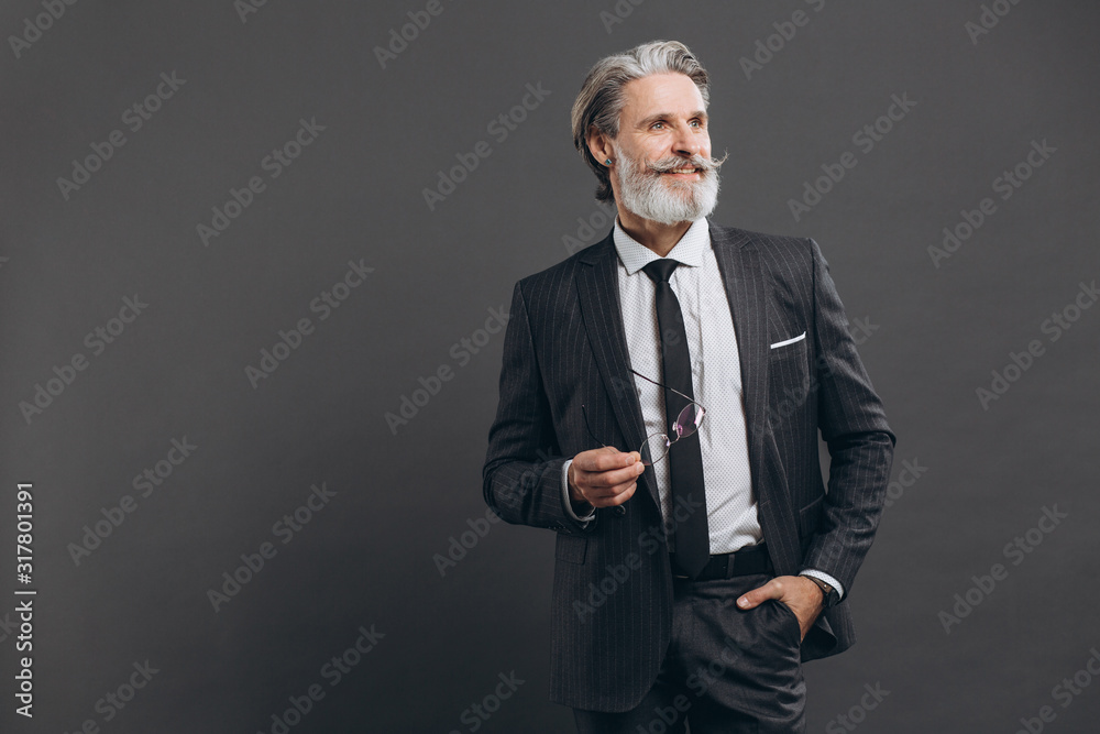 Business and fashionable bearded mature man in a gray suit holding his glasses and smiling at the copy space on a grey background. Business and fashionable bearded mature man in a gray suit holding his glasses and smiling at the copy space on a grey background.
