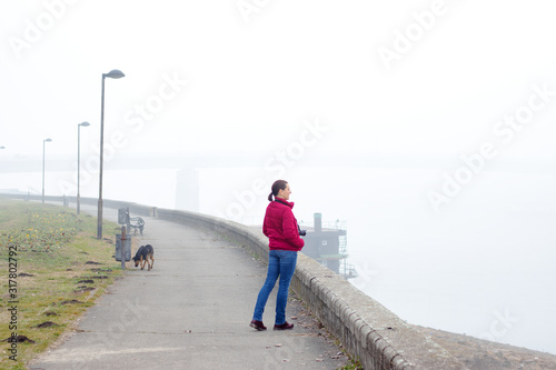 Young woman standing at river bank  with camera hanging around her neck  looking through thick fog for the other side of the river. Conceptual photo about ecology and air pollution.
