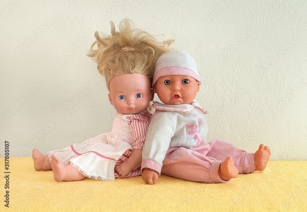 Tiny tots dolls in pink clothes. Nursery dolls sitting back to back on yellow underlay next to the wall. Caucasian newborn baby puppets. Studio shot. Pair of figureheads sitting behind another. Tiny tots dolls in pink clothes. Nursery dolls sitting back to back on yellow underlay next to the wall. Caucasian newborn baby puppets. Studio shot. Pair of figureheads sitting behind another.