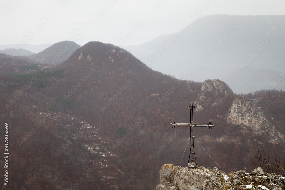 Large metallic cross on a edge of a mountain cliff and impressive background mountains covered by snow, autumn colored trees and fog
