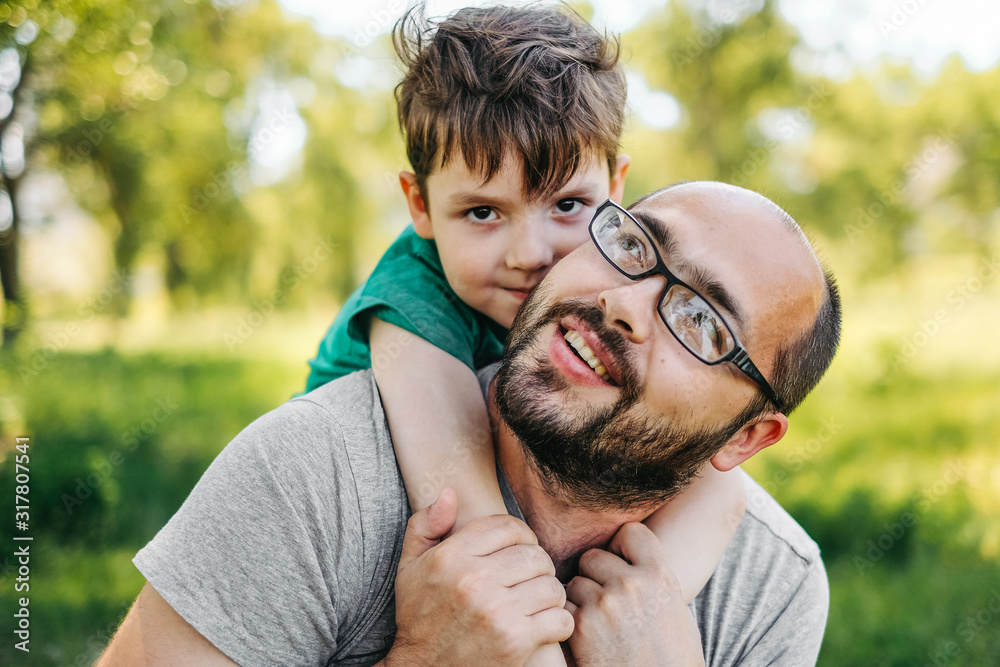 Fototapeta premium Portrait of handsome dad with a beard, looking at camera and smiling, hugging his cute little boy, trees in the background