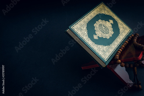 Holy Al Quran with written arabic calligraphy meaning of Al Quran and tasbih or rosary beads on black background. Selective focus and crop fragment