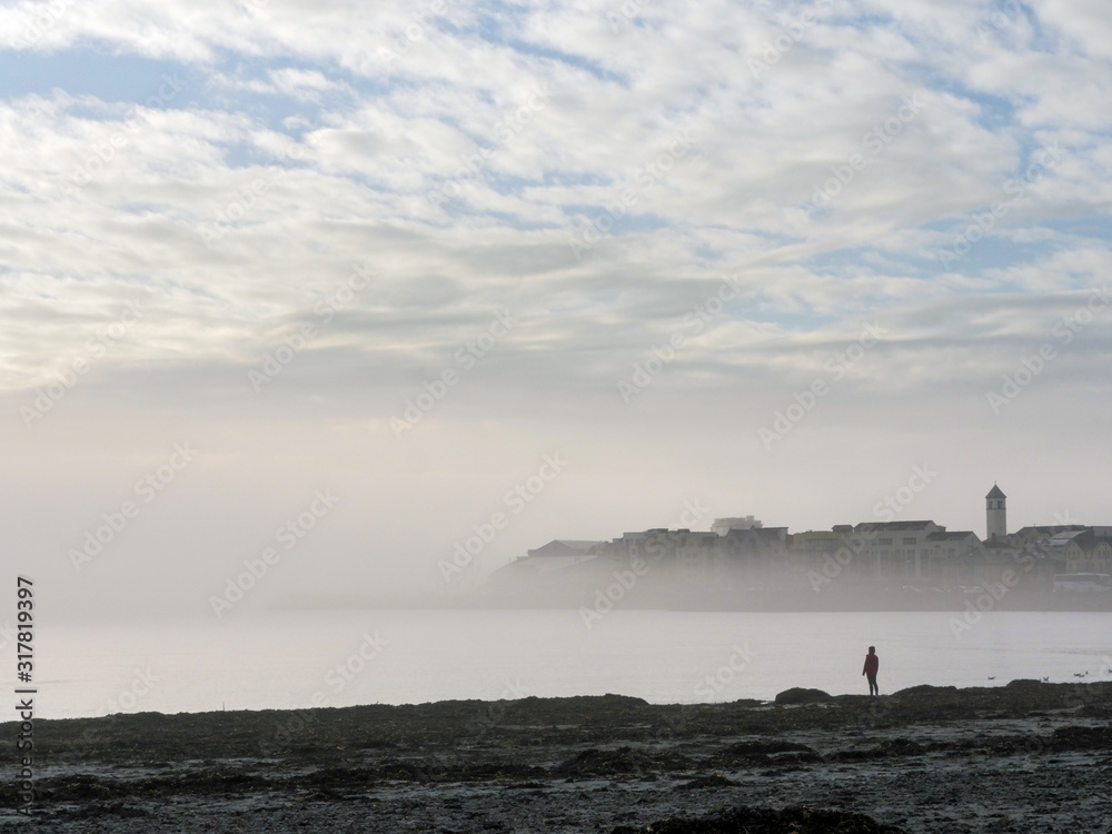 Galway bay and Grattan beach, Cloudy sky, Fog over houses, one person silhouette standing by the ocean, low tide.