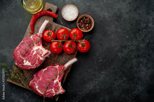 two raw steaks, beef tomahawk with spices, tomatoes and thyme on a stone background. with copy space for your text