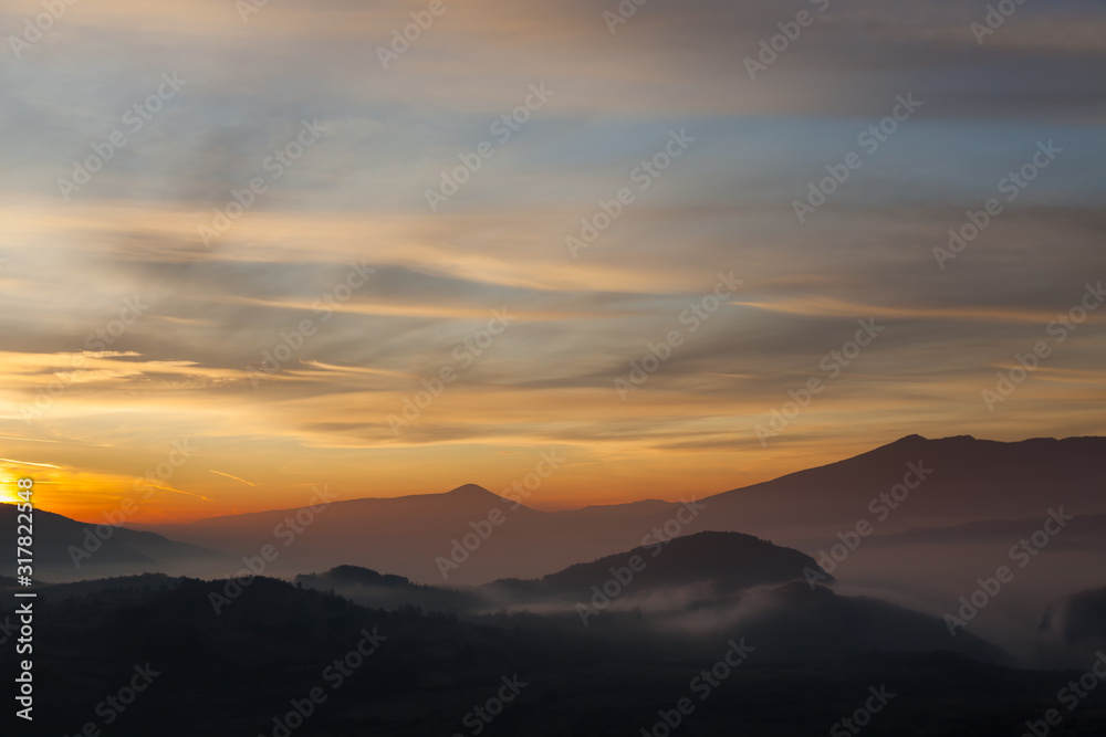 Valley filled with soft mist, distant silhouettes of impressive mountains and amazing colors of vivid morning sky