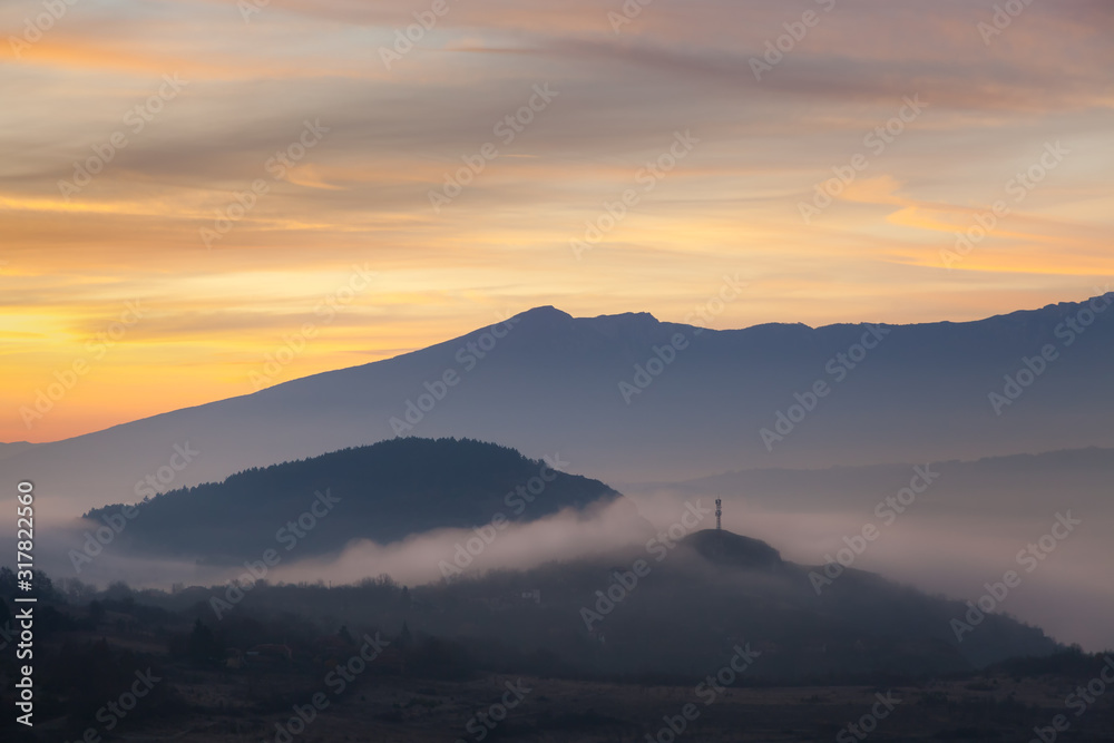 Soft, morning mist covering village houses, mountain layers and valley, distant pointy mountain peak and amazing vivid colors of sunrise sky