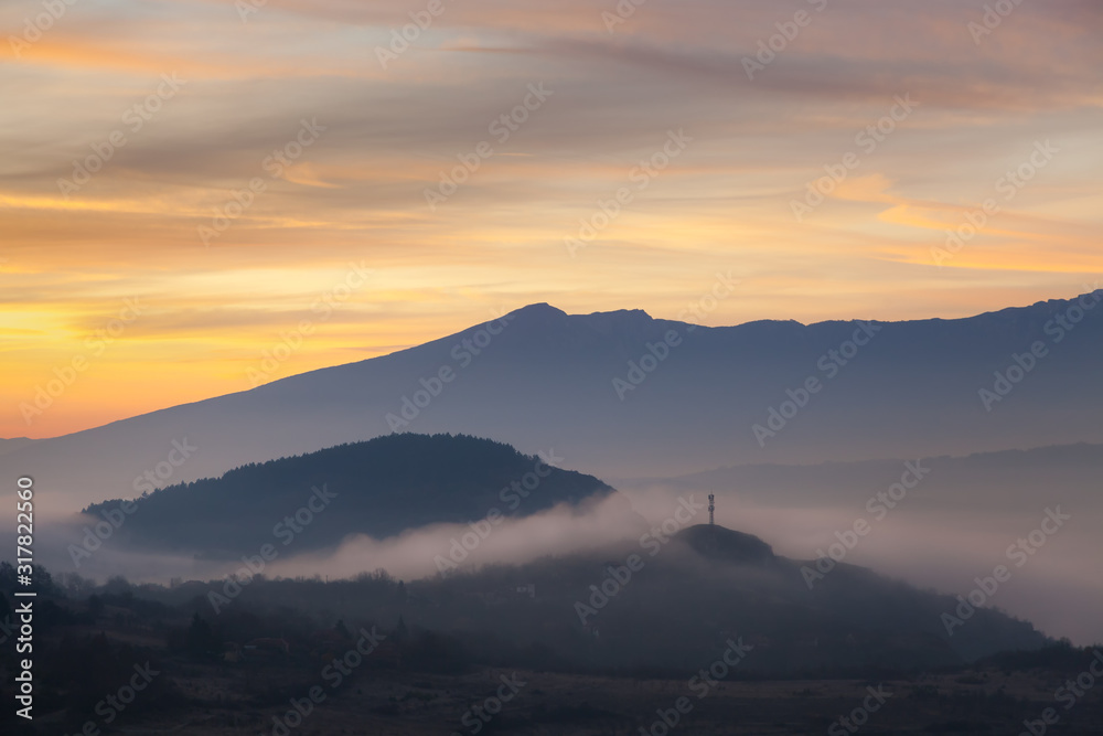 Soft, morning mist covering village houses, mountain layers and valley, distant pointy mountain peak and amazing vivid colors of sunrise sky Soft, morning mist covering village houses, mountain layers and valley, distant pointy mountain peak and amazing vivid colors of sunrise sky