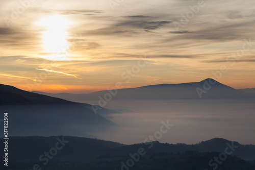 Valley filled with soft mist, distant silhouettes of impressive mountains and amazing colors of vivid morning sky