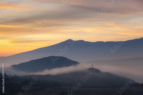 Soft  morning mist covering village houses  mountain layers and valley  distant pointy mountain peak and amazing vivid colors of sunrise sky