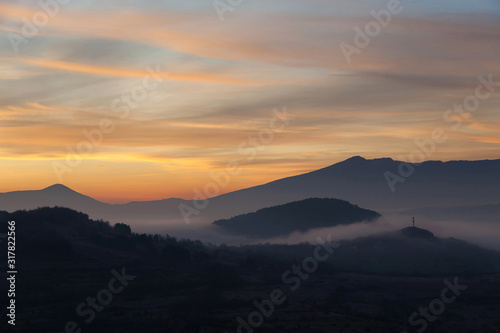 Soft  morning mist covering village houses  mountain layers and valley  distant pointy mountain peak and amazing vivid colors of sunrise sky