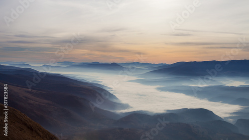Soft, abstract, moody view of a valley filled with thick morning fog, colorful sunrise sky and impressive mountains © Nikola