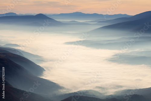 Soft, abstract, moody view of a valley filled with thick morning fog, colorful sunrise sky and impressive mountains © Nikola