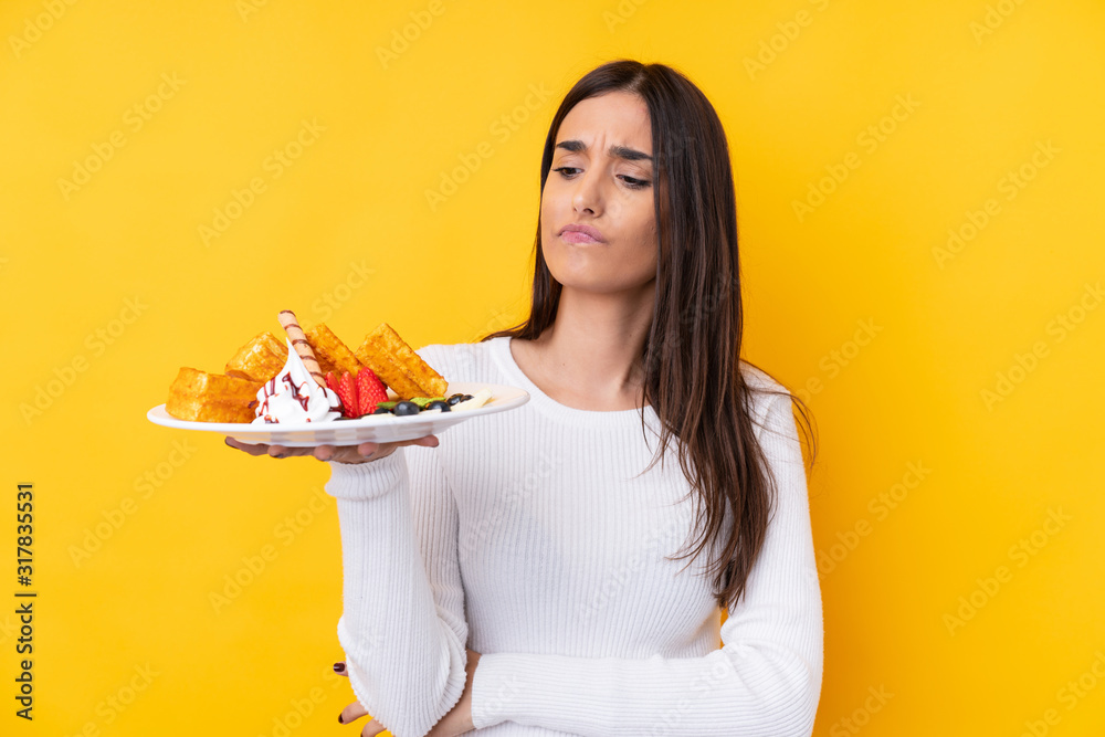 Young brunette woman holding waffles over isolated background with sad expression Young brunette woman holding waffles over isolated background with sad expression