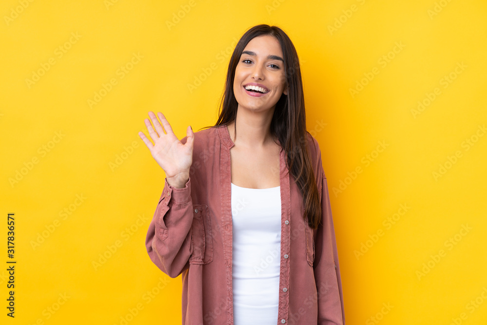 Young brunette woman over isolated yellow background saluting with hand with happy expression Young brunette woman over isolated yellow background saluting with hand with happy expression