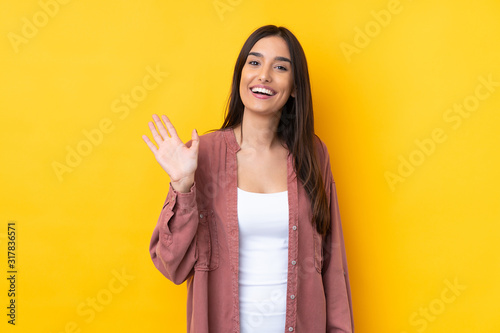 Young brunette woman over isolated yellow background saluting with hand with happy expression