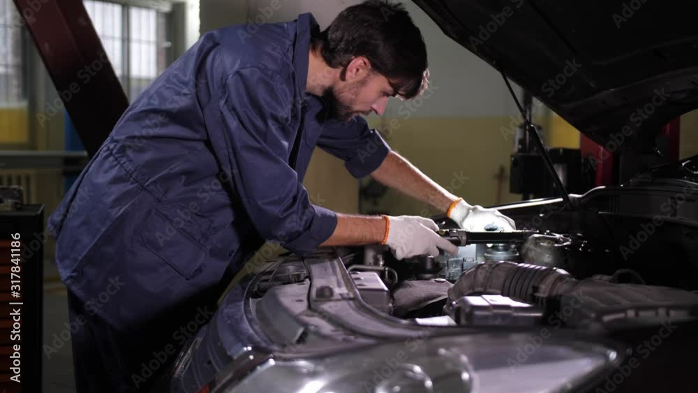 Closeup of auto repair service worker taking flashlight to check brake and coolant levels during vehicle maintenance. Handsome repairman standing near opened car hood and highlighting space under hood Closeup of auto repair service worker taking flashlight to check brake and coolant levels during vehicle maintenance. Handsome repairman standing near opened car hood and highlighting space under hood