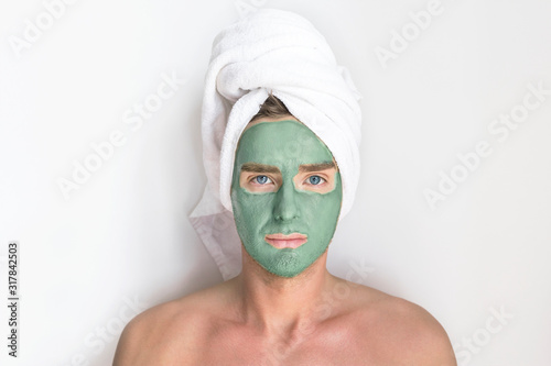 Young handsome man, metrosexual guy with cosmetic clay blue mask on his face and a towel on head looking at camera, smiling on white background. Beauty, spa, skin care concept. Men taking care of skin