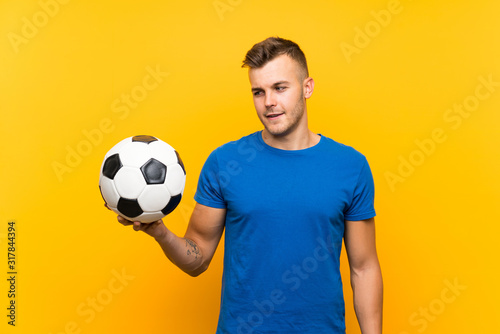 Young handsome blonde man holding a soccer ball over isolated yellow background with happy expression