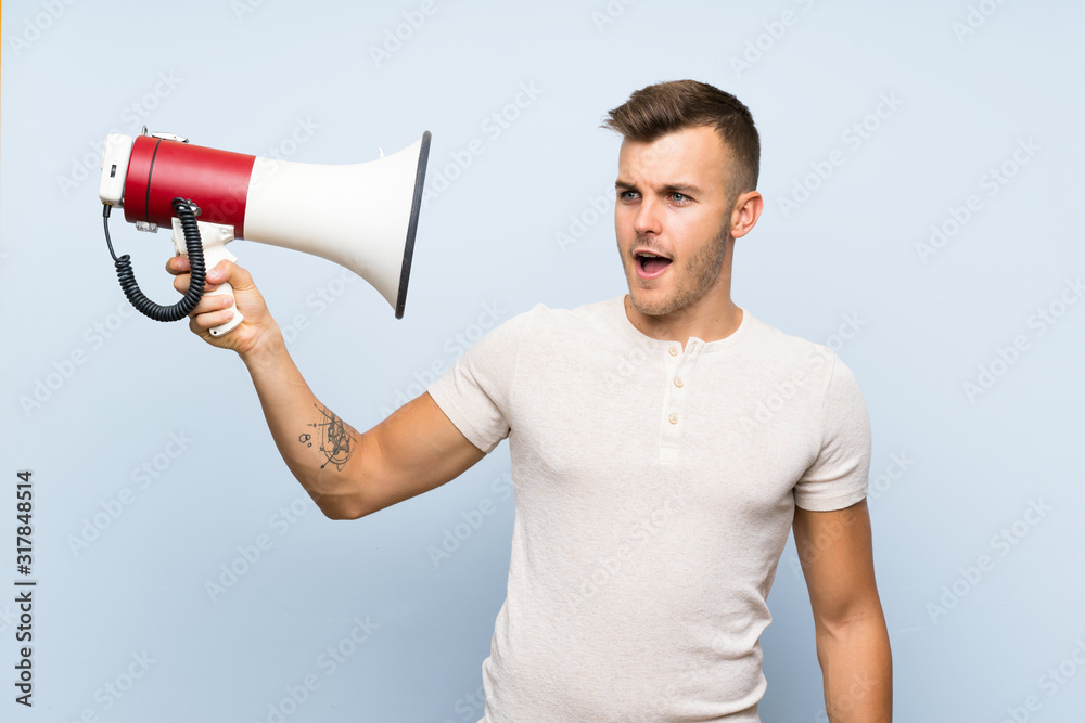 Young handsome blonde man over isolated blue background holding a megaphone Young handsome blonde man over isolated blue background holding a megaphone